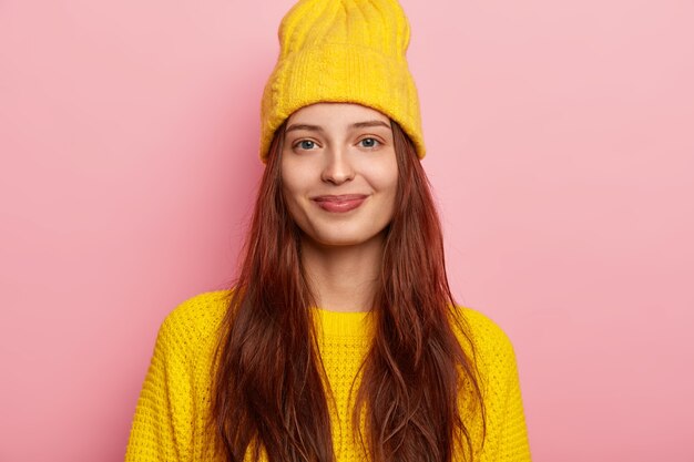 Das Bild des zufriedenen reizenden jungen weiblichen Modells im stilvollen gelben Hut und im gestrickten Pullover, hat langes Haar, posiert vor rosa Hintergrund, zeigt ihre Winterkleidung, die direkt mit leichtem Lächeln in die Kamera schaut