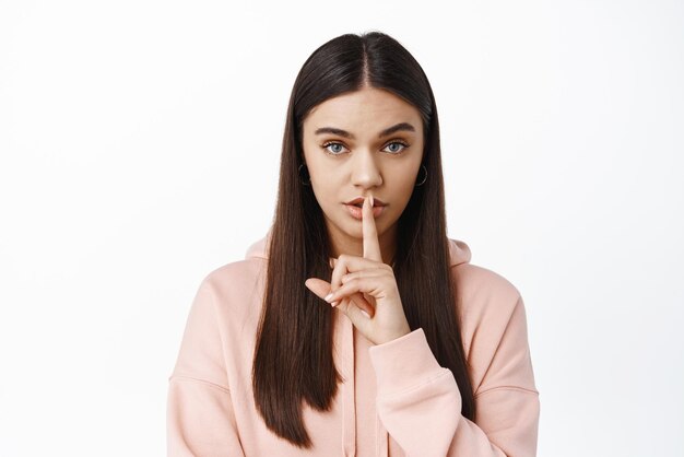 Das Bild einer ernsthaften Frau macht eine Tabu-Geste, zeigt ein Psst-Psst-Zeichen auf den Lippen und starrt in die Kamera und bittet um ein geheimes Stehen im Hoodie vor weißem Hintergrund
