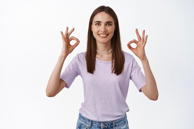 Das Bild einer lächelnden jungen Frau zeigt OK-Zeichen, keine Probleme, empfiehlt etwas Gutes, positives Feedback, mag und stimmt zu, genehmigt, steht auf weißem Hintergrund