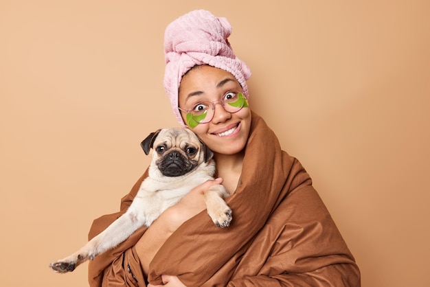 Das Bild einer positiven jungen Frau hält einen Pug-Hund trägt ein gewickeltes Handtuch auf dem Kopf und eine weiche Decke posiert vor braunem Hintergrund und drückt die Liebe zum Haustier aus Fröhlicher Haustierbesitzer mit Lieblingshaustier