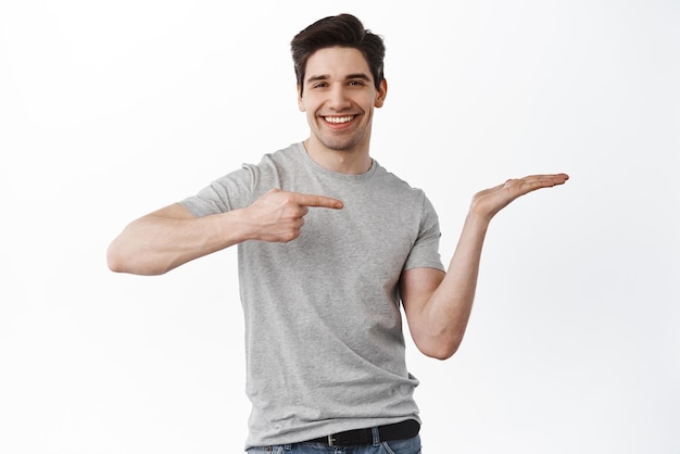 Das Bild eines gutaussehenden lächelnden Mannes, der auf seine leere Hand zeigt, die einen Gegenstand auf der Handfläche hält, zeigt Ihr Produkt und zeigt den Kopienraum, der zufrieden vor weißem Hintergrund steht