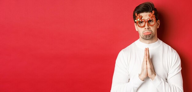 Das Bild eines gutaussehenden Mannes in einer Weihnachtsfeierbrille, der um Hilfe bittet oder sich entschuldigt, braucht einen Gefallen und steht auf rotem Hintergrund.