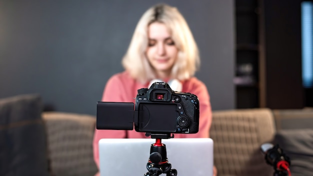 Das blonde Mädchen des jungen Inhaltsschöpfers ist auf ihrem Laptop auf dem Tisch. Sich mit einer Kamera auf einem Stativ filmen. Von zu Hause aus arbeiten. Vlog aufzeichnen