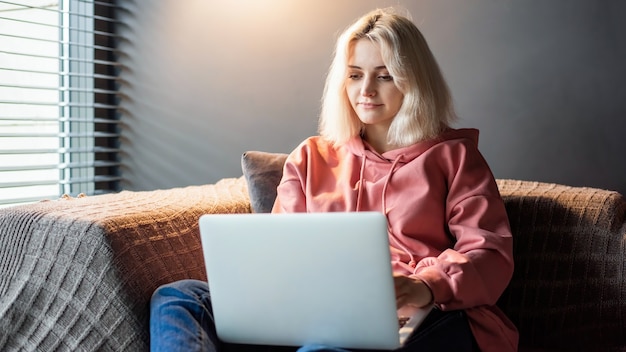 Das blonde Mädchen des jungen Inhaltsschöpfers sitzt auf ihrem Laptop auf dem Sofa in der Nähe des Fensters. Von zu Hause aus arbeiten