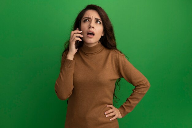 Das Denken, das junges schönes Mädchen nach oben schaut, spricht am Telefon, das Hand auf Hüfte lokalisiert auf grüner Wand setzt