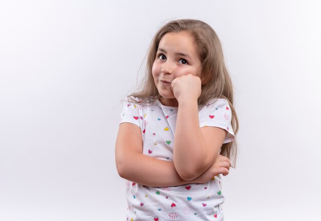 Das denkende kleine Schulmädchen, das weißes T-Shirt trägt, legte ihre Faust auf Wange auf isolierte weiße Wand