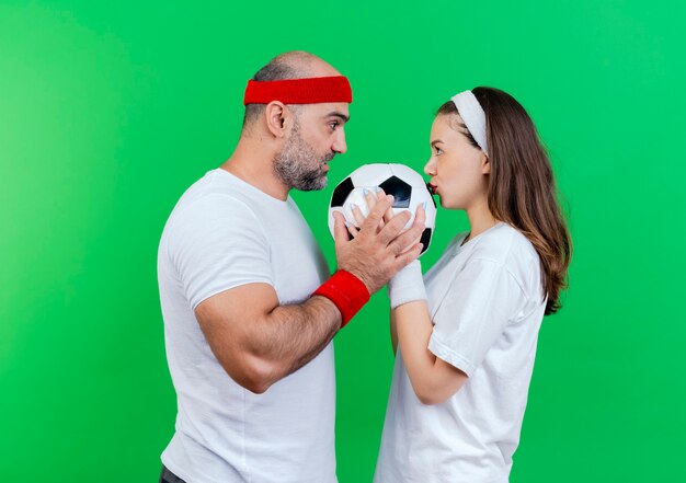 Das erwachsene sportliche paar, das stirnband und armbänder trägt, beeindruckte mann, der fußballfußballfrau hält, die ball küsst, der einander ansieht