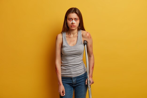 Das Foto einer Frau, die Opfer eines Unfalls ist, hat einen Nasenbruch, posiert mit einer Krücke, kann nicht alleine gehen, bekommt die Folgen eines unachtsamen Fahrens, trägt eine Weste, Jeans, hat Abrieb und blaue Flecken auf der Haut