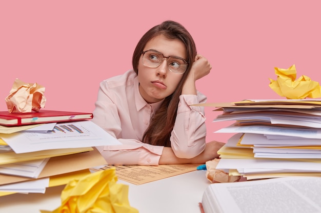 Das Foto einer müden Frau hat keine Lust zu arbeiten, schaut nachdenklich zur Seite, trägt eine große Brille, hat viel Papier zum Durchschauen, sitzt alleine am Arbeitsplatz