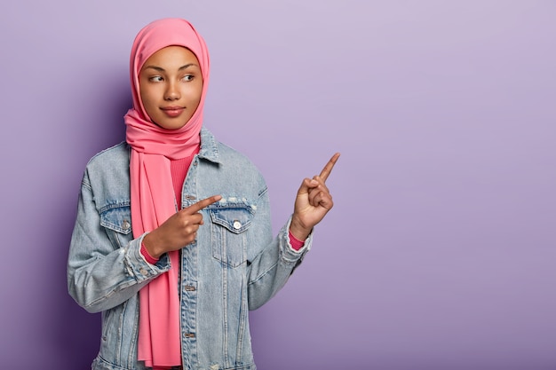 Das Foto einer selbstbewussten arabischen Dame in traditionellem rosa Hijab zeigt Ihnen den richtigen Weg, zeigt mit beiden Vorderfingern auf eine leere Stelle, lädt ein, dorthin zu gehen, fördert den Kopierraum. Probieren Sie es mit mir aus