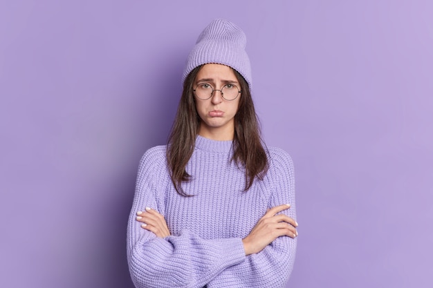 Das foto einer unzufriedenen brünetten frau sieht mit beleidigtem ausdruck aus und drückt die hände, wenn jemand verletzt wird. er trägt eine runde brille und lila kleidung. mürrische frau hat schlechte laune. ressentimentskonzept