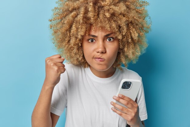 Das Foto einer wütenden, empörten, lockigen Frau, die die Lippen mit der Faust ballt, sieht wütend aus und verwendet ein modernes Mobiltelefon, um Textnachrichten zu senden, die in einem lässigen weißen T-Shirt gekleidet sind, das über blauem Hintergrund isoliert ist