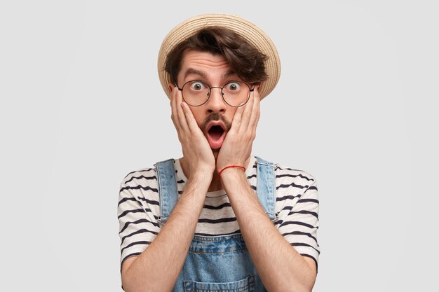 Das Foto eines betäubten jungen bärtigen männlichen Bauern hält beide Handflächen auf den Wangen, hört schlechte Nachrichten über die Situation auf dem Bauernhof, gekleidet in ein weiß gestreiftes T-Shirt und Jeans-Overllas, isoliert über einer weißen Wand