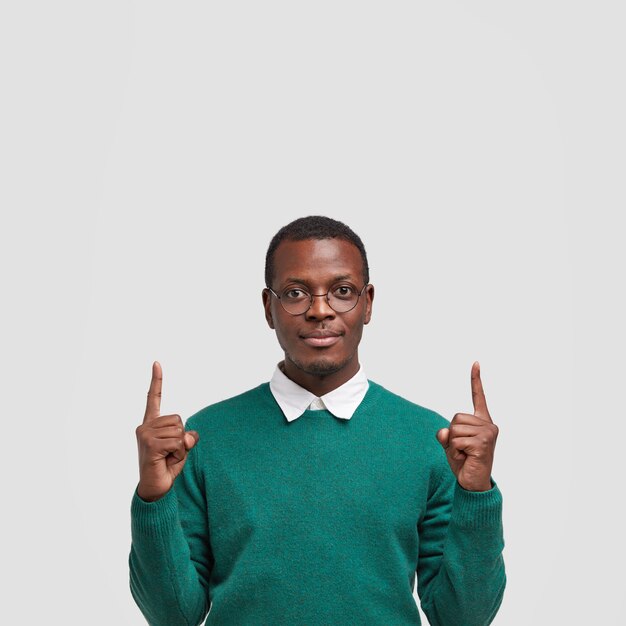 Das Foto eines ernsthaften dunkelhäutigen Mannes mit selbstbewusstem Gesichtsausdruck zeigt mit beiden Zeigefingern nach oben, trägt eine Brille und einen grünen Pullover