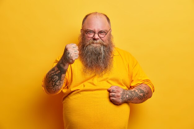 Das Foto eines ernsthaften wütenden Mannes hat einen dicken Bart, ballt die Fäuste und sieht mit empörtem Ausdruck aus, verspricht Rache, zeigt einen dicken dicken Bauch, gekleidet in ein gelbes T-Shirt, drückt negative Gefühle aus