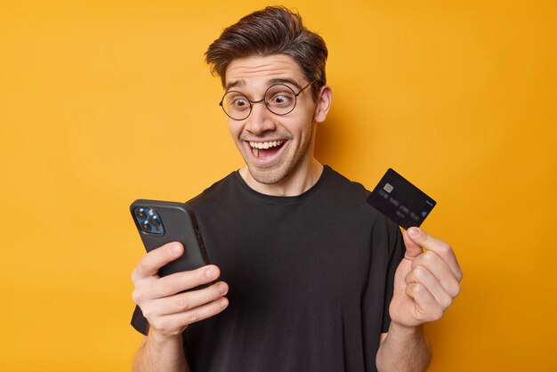 Das Foto eines fröhlichen, überraschten erwachsenen Mannes starrt mit einem unglaublichen Blick auf das Smartphone, der schockiert ist, eine große Menge Geld zu erhalten, trägt eine Brille und ein lässiges schwarzes T-Shirt, das auf gelbem Hintergrund isoliert ist