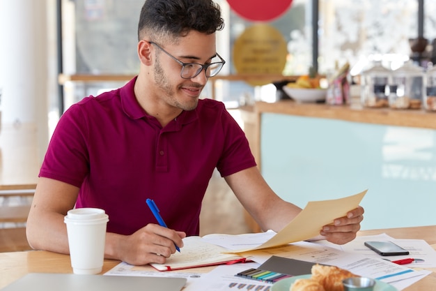 Das Foto eines jungen unerfahrenen Unternehmers zeichnet Informationen aus Geschäftsdokumenten im Notizblock auf, studiert Grafiken und Diagramme, bereitet sich auf die Präsentation von Informationen für Investoren vor und trinkt Kaffee