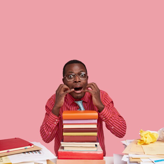 Das Foto eines nervösen schwarzen Mannes hält die Hände in der Nähe des Mundes, starrt mit angstvollen Augen, hat einen Stapel Lehrbücher auf dem Schreibtisch, Papiere und Antworten auf die Prüfung