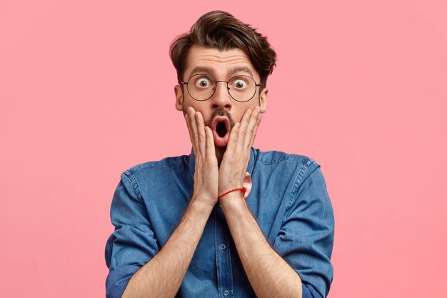 Das Foto eines verblüfften bärtigen Mannes mit stilvollem Haarschnitt hält die Hände auf beiden Wangen, sieht überraschend und schockiert aus, öffnet den Mund weit, trägt ein Jeanshemd und posiert an der rosa Wand