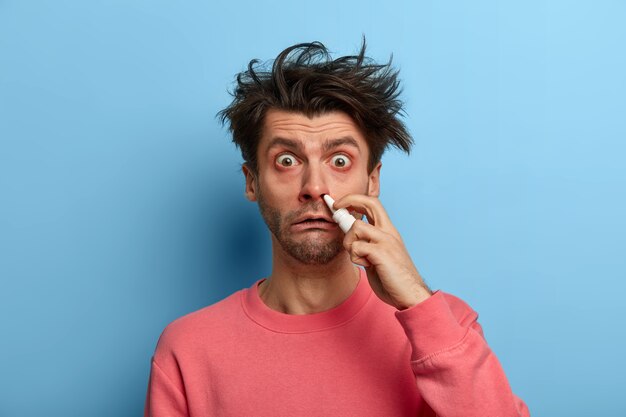 Das Foto eines verlegenen kranken Mannes hat die Nase blockiert, verwendet wirksame Medikamente, hält eine Flasche Nasentropfen zum Atmen frei, trägt einen rosa Pullover und wirbt für Mittel gegen laufende Nase. Menschen, Erkältung, Behandlung
