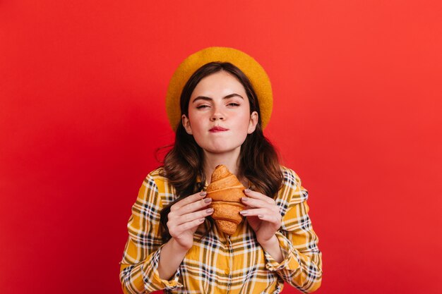 Das freche dunkelhaarige Mädchen beißt sich auf die Lippe und möchte leckeres Croissant essen. Dame im Hut und in der gelben Bluse, die auf roter Wand aufwirft.