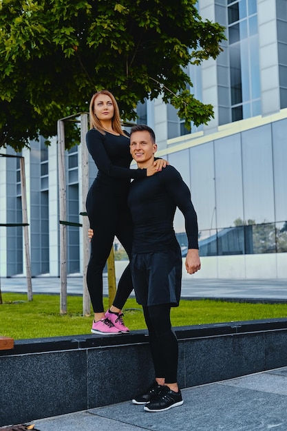 Das ganzkörperbild eines athletischen fitnesspaares posiert vor einem modernen gebäudehintergrund.