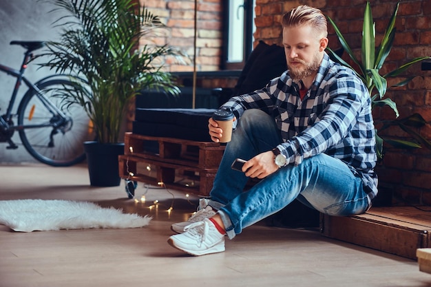 Das Ganzkörperbild eines blonden Hipster-Männchens trinkt Kaffee in einem Raum mit Loft-Interieur.