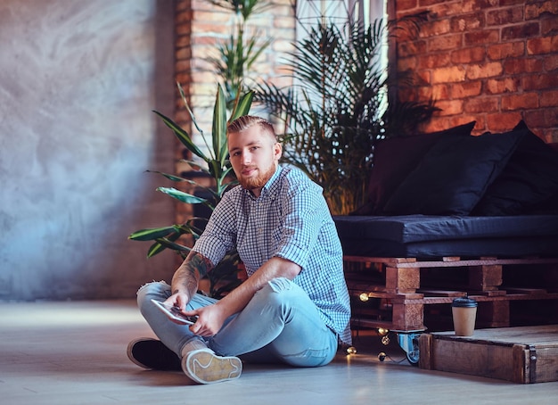 Das Ganzkörperbild eines blonden, stylischen Mannes in Fleecehemd und Jeans mit einem Tablet-PC in einem Wohnzimmer.