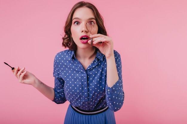 Das geschockte weiße Mädchen in der trendigen Bluse kräuselt ihre Wimpern an der rosa Wand. spektakuläre verblüffte Frau mit dunklem Haar bereitet sich auf Date vor.