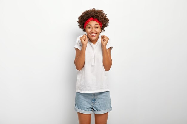 Das glückliche afroamerikanische Mädchen ballt die Fäuste, glaubt an den Sieg, lächelt fröhlich, zeigt weiße Zähne, trägt Freizeitkleidung, posiert drinnen, fühlt sich wie ein Gewinner, jubelt allein