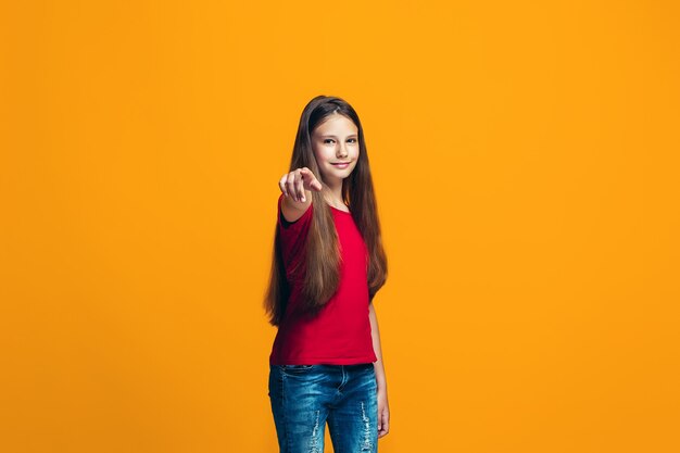 Das glückliche jugendlich Mädchen, das auf Sie zeigt, halbes Länge Nahaufnahmeporträt auf orange Hintergrund.