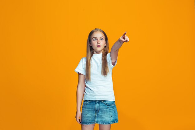 Das glückliche jugendlich Mädchen zeigt auf Sie, halbes Länge Nahaufnahmeporträt auf orange Raum.
