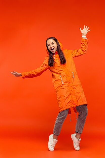 Das glückliche lächelnde junge Mädchen, das im Studio in der orangefarbenen Herbstjacke auf Rot aufwirft. Menschliche positive Emotionen. Konzept des kalten Wetters. Weibliche Modekonzepte