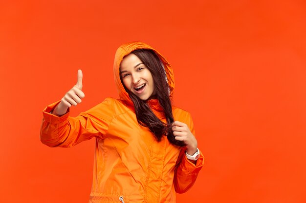 Das glückliche lächelnde junge Mädchen, das im Studio in der orangefarbenen Herbstjacke auf Rot aufwirft.