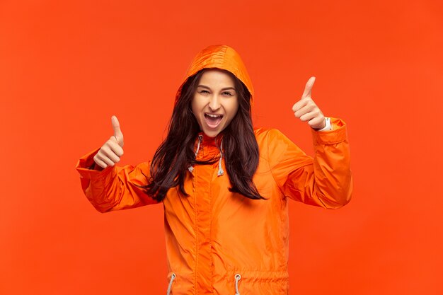 Das glückliche lächelnde junge Mädchen, das im Studio in der orangefarbenen Herbstjacke auf Rot aufwirft.