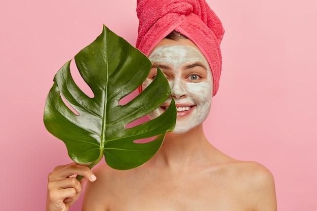 Das glückliche weibliche Model hat eine gründliche Reinigung mit Gesichtsmaske, bedeckt die Hälfte des Gesichts mit grünem Blatt, verbessert ihr Aussehen, möchte eine fabelhafte Haut haben, verstopft die Poren und lächelt sanft