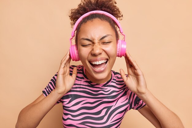 Das horizontale Bild der überglücklichen jungen Frau lächelt im Großen und Ganzen genießt eine angenehme Melodie hält die Hände auf den Kopfhörern hat eine optimistische Stimmung in gestreiften rosa und schwarzen T-Shirt-Posen im Innenbereich