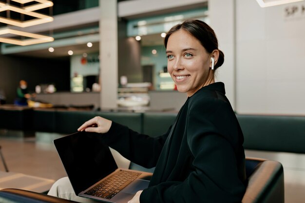 Das Innenporträt einer liebenswerten süßen Dame mit dunklem Haar trägt einen dunklen Anzug in Kopfhörern und blickt mit einem glücklichen Lächeln in die Kamera, während sie im modernen Büro arbeitet