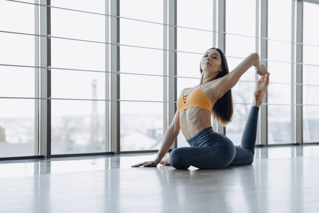 Das junge attraktive Mädchen, das Eignung tut, trainiert mit Yoga auf dem Boden von panoramischen Fenstern