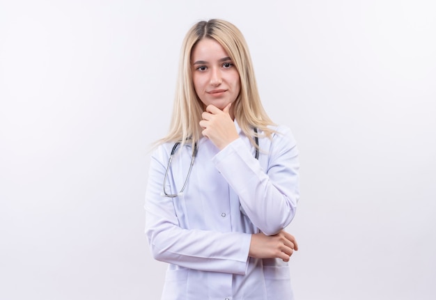 Das junge blonde Mädchen des Doktors, das Stethoskop und medizinisches Kleid trägt, legte ihre Hand auf Kinn auf isolierte weiße Wand