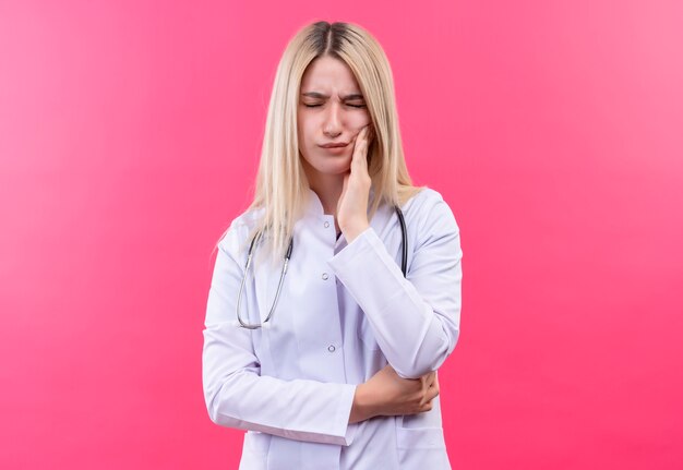 Das junge blonde Mädchen des kranken Arztes, das Stethoskop im medizinischen Kleid trägt, legte ihre Hand auf schmerzenden Zahn auf lokalisiertem rosa Hintergrund