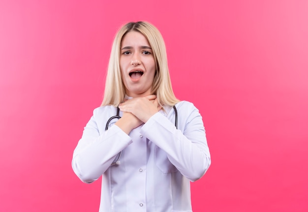 Das junge blonde Mädchen des kranken Arztes, das Stethoskop im medizinischen Kleid trägt, packte die Kehle mit beiden Händen auf lokalisiertem rosa Hintergrund