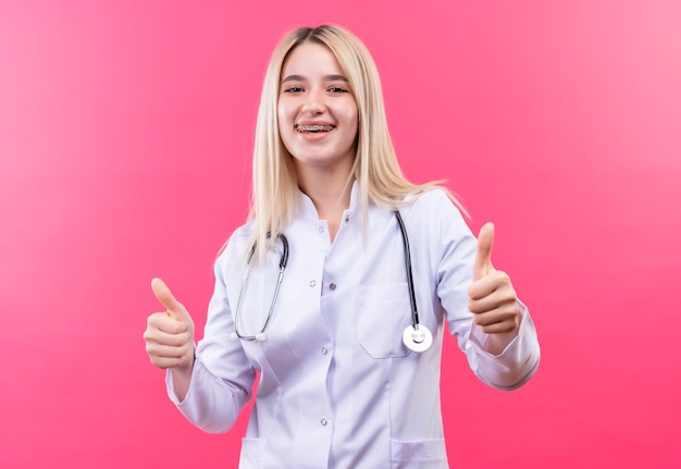 Das junge blonde Mädchen des lächelnden Doktors, das Stethoskop im medizinischen Kleid und in der Zahnspange trägt, ihre Daumen oben auf lokalisiertem rosa Hintergrund