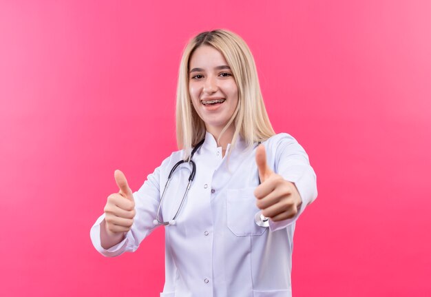 Das junge blonde Mädchen des lächelnden Doktors, das Stethoskop im medizinischen Kleid und in der Zahnspange trägt, ihre Daumen oben auf lokalisiertem rosa Hintergrund