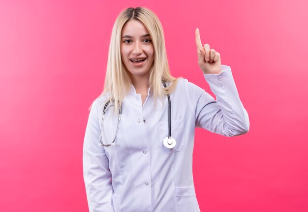 Das junge blonde Mädchen des lächelnden Doktors, das Stethoskop im medizinischen Kleid und in der Zahnspange trägt, zeigt auf lokalisierten rosa Hintergrund