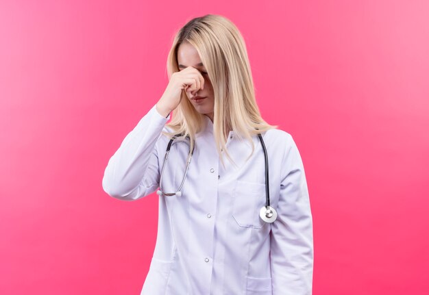 Das junge blonde Mädchen des müden Arztes, das Stethoskop im medizinischen Kleid trägt, legte ihre Hand auf Nase auf isolierte rosa Wand