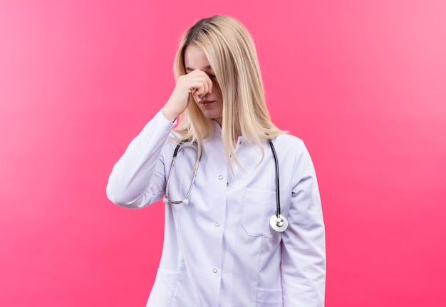 Das junge blonde Mädchen des müden Arztes, das Stethoskop im medizinischen Kleid trägt, legte ihre Hand auf Nase auf isolierte rosa Wand
