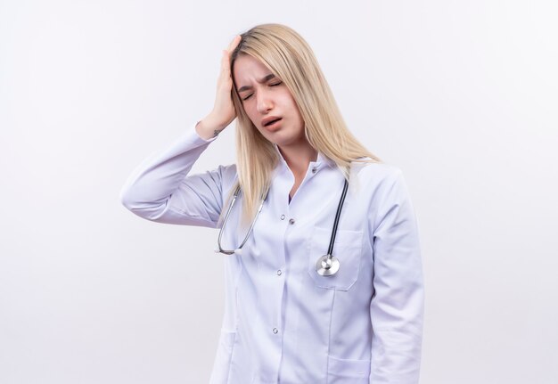 Das junge blonde Mädchen des müden Arztes, das Stethoskop und medizinisches Kleid trägt, legte ihre Hand auf Kopf auf isolierte weiße Wand