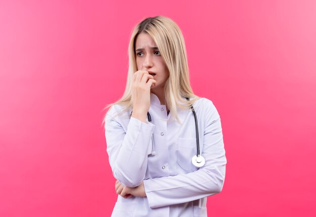 Das junge blonde Mädchen des traurigen Doktors, das Stethoskop im medizinischen Kleid trägt, legte ihre Hand auf Kinn auf lokalisiertem rosa Hintergrund