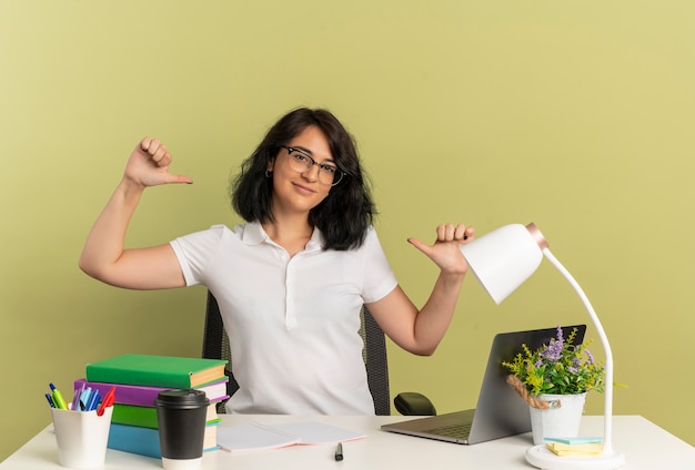 Das junge erfreute hübsche kaukasische Schulmädchen, das eine Brille trägt, sitzt am Schreibtisch mit den Schulwerkzeugpunkten auf sich selbst isoliert auf Grünfläche mit Kopienraum